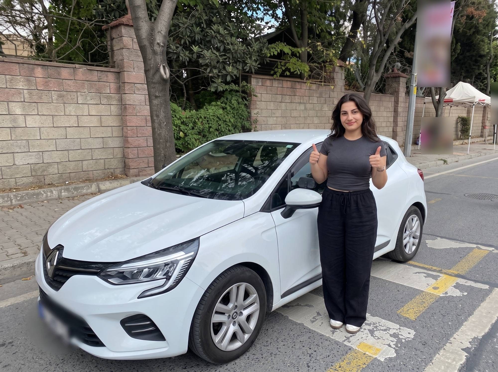 Beyaz bir otomobilin yanında başparmaklarını kaldırarak gülümseyen öğrenci